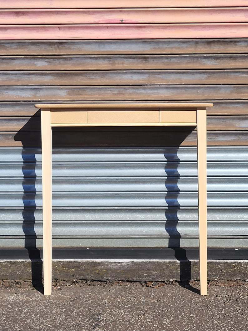 May include: A light-colored wooden console table featuring a rectangular top and four straight, slender legs. The front of the table includes a horizontal panel with three distinct sections, suggesting the presence of drawers or storage compartments.