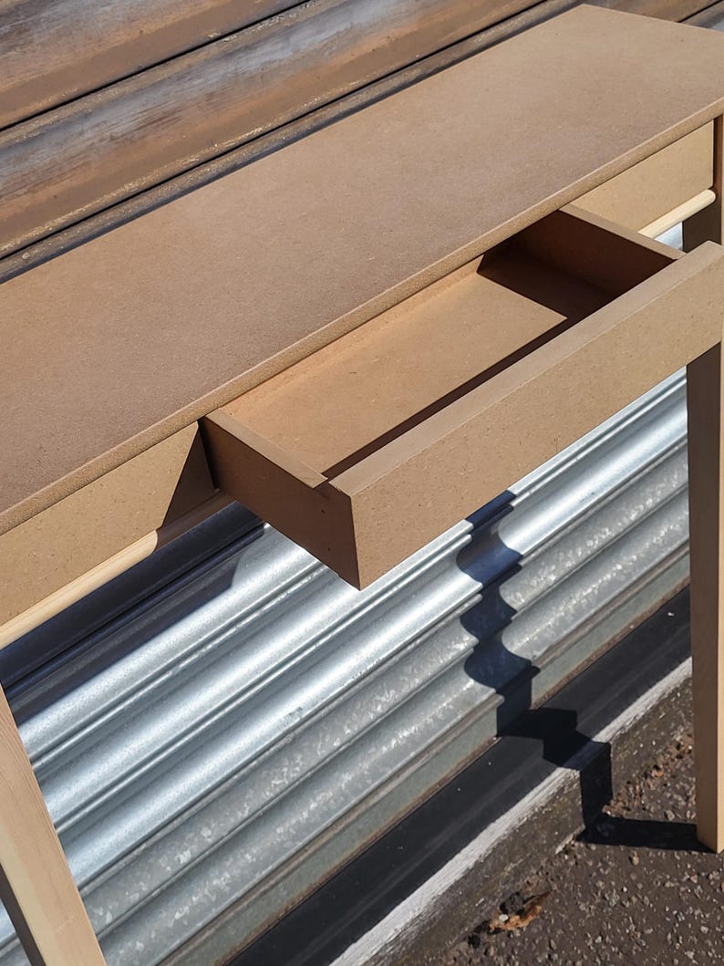 May include: A light brown console table constructed from unfinished medium-density fiberboard. The table features a flat rectangular top surface and a single pull-out drawer positioned centrally beneath the top, showcasing a smooth, matte texture.