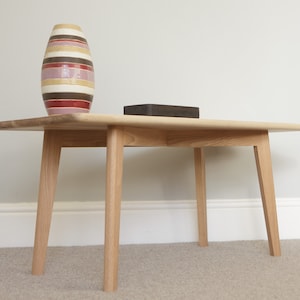 May include: A light wood coffee table with four legs. The table has a rectangular top and a slightly rounded edge. A vase with brown, pink, and white stripes sits on the table.