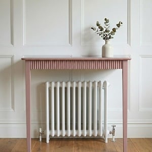 May include: A dusty pink console table with a fluted apron design and four straight legs. The table features a flat rectangular top surface. A small, off-white ceramic vase containing a sprig of green eucalyptus leaves sits on the table top.
