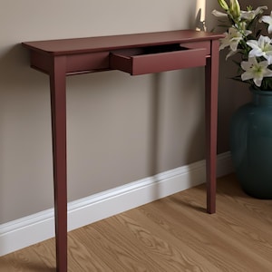May include: A slim, rectangular console table finished in a deep matte burgundy color. The table features four straight, slender legs and a single, centrally positioned drawer that is pulled outward, revealing its open, hollow interior space.