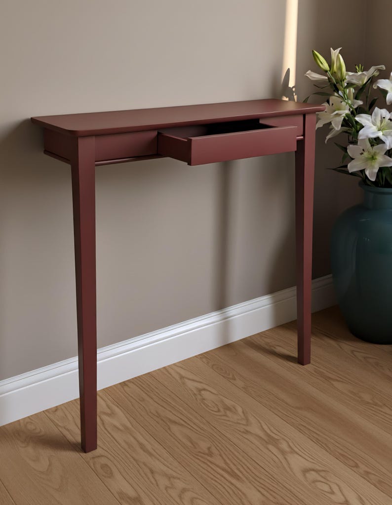 May include: A slim, rectangular console table finished in a deep matte burgundy color. The table features four straight, slender legs and a single, centrally positioned drawer that is pulled outward, revealing its open, hollow interior space.