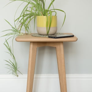 May include: A small wooden end table with a light wood finish and three legs. A potted plant with green leaves is on the table, and a black smartphone is on the table next to the plant.