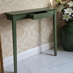 May include: A sage green console table featuring a rectangular top, four straight legs, and a single central drawer that is partially pulled open. The furniture piece has a smooth, matte finish and a clean, linear design with no visible hardware.