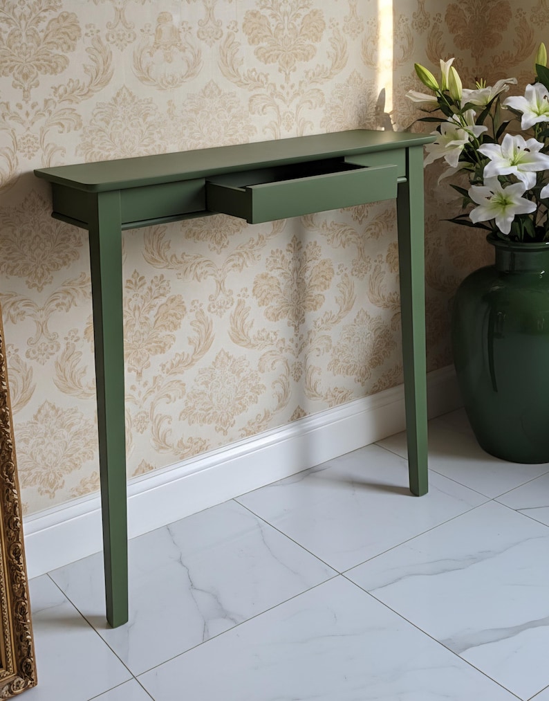 May include: A sage green console table featuring a rectangular top, four straight legs, and a single central drawer that is partially pulled open. The furniture piece has a smooth, matte finish and a clean, linear design with no visible hardware.