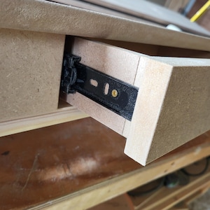 May include: A light brown medium-density fiberboard drawer partially extended from a matching frame. A black metal ball-bearing drawer slide is visible on the side, secured with a single gold-colored screw through an elongated mounting slot.