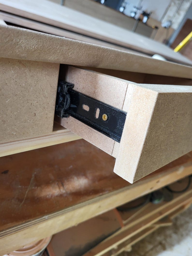 May include: A light brown medium-density fiberboard drawer partially extended from a matching frame. A black metal ball-bearing drawer slide is visible on the side, secured with a single gold-colored screw through an elongated mounting slot.