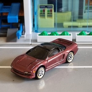 Puede incluir: Un coche de juguete de metal fundido a presión en color granate metalizado con techo negro, ventanas tintadas y ruedas con llantas blancas. El vehículo presenta un diseño aerodinámico de perfil bajo con faros empotrados y un alerón trasero.