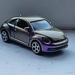 Puede incluir: Un modelo de coche de juguete de un Volkswagen Beetle en color verde oscuro y morado con un acabado brillante. El coche es una versión moderna del Beetle clásico.