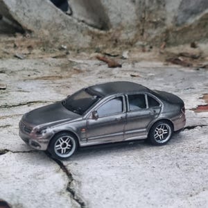 Könnte beinhalten: Ein silbernes Spielzeug-Limousinenauto mit schwarz-silbernen Rädern. Das Miniaturfahrzeug steht auf einer strukturierten Oberfläche. Das Auto hat ein schlankes Design mit sichtbaren Fenstern und Seitenspiegeln. Das Spielzeugauto ist ein Sammlerstück.