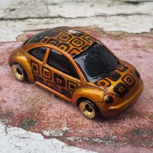 Pode incluir: Um carro de brinquedo dourado com um padrão geométrico preto e laranja. O carro é um Volkswagen Beetle.