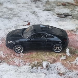 Puede incluir: Un coche de juguete negro con llantas plateadas se encuentra sobre una superficie nevada. El vehículo en miniatura tiene un diseño elegante y un acabado brillante. Las ruedas del coche son detalladas, y la estética general sugiere un coche deportivo.