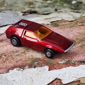 Puede incluir: Un pequeño coche de juguete rojo con un diseño aerodinámico. El coche tiene un parabrisas amarillo y ruedas negras. Este coche de juguete es un objeto de colección vintage, probablemente de los años 70 u 80.
