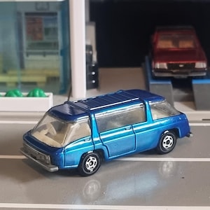 Puede incluir: Una furgoneta de juguete azul fundido a presión con ventanas transparentes y ruedas negras. La furgoneta de juguete está estacionada sobre una superficie gris, con un garaje en miniatura y un coche rojo en el fondo. La furgoneta tiene un diseño detallado.