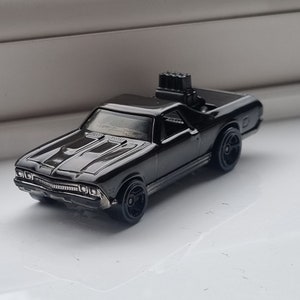 Könnte beinhalten: Ein schwarzes Spielzeugautomodell eines Chevrolet El Camino Pickups mit einem schwarzen Auspuff auf der Ladefläche. Das Auto hat schwarze Räder und eine schwarze Karosserie.