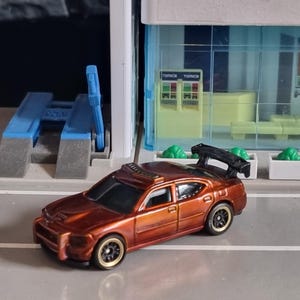 Puede incluir: Un coche de juguete de color bronce con ruedas negras y un alerón trasero. El coche tiene un diseño detallado, incluyendo una barra de luces en el techo. El fondo presenta un escenario de garaje de juguete.