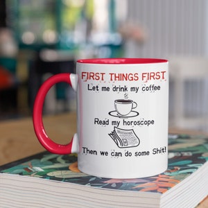 May include: White ceramic coffee mug with a red rim. The mug has black text that reads "FIRST THINGS FIRST Let me drink my coffee Read my horoscope Then we can do some Shit!" with a small illustration of a cup of coffee and a newspaper with the word "Horoscope" on it.