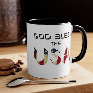 Peut inclure: Mug en céramique blanche avec intérieur et anse noirs. Le mug affiche le texte "GOD BLESS THE USA" en noir, avec les lettres "USA" conçues avec un motif de drapeau américain vieilli. Une cuillère et un macaron sont au premier plan.