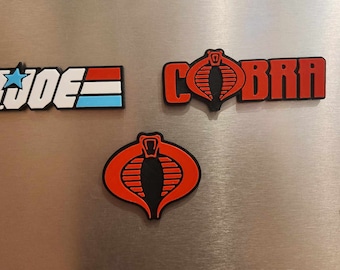 May include: Three refrigerator magnets featuring the logos of G.I. Joe and Cobra. The G.I. Joe magnet is white with a blue and red stripe and a star. The Cobra magnets are red with a black cobra logo and the word "Cobra" in black.