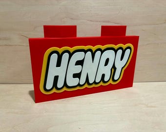 May include: A red rectangular sign shaped like a building block with two studs on top. The center features the name HENRY in white letters with a thick black outline, set against a yellow background that follows the contour of the text.