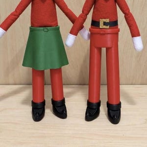 May include: Two figures with red felt long-sleeved tops and white cuffs. One figure wears a green skirt, the other wears red trousers with a black belt and gold buckle. Both figures have black pointed shoes and stand upright on a light wood surface.