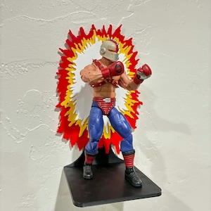 Peut inclure: Figurine d'action musclée en position de combat, avec un casque, des gants rouges et un pantalon bleu. Elle est présentée sur une étagère noire avec un fond en forme d'explosion rouge, jaune et blanc.