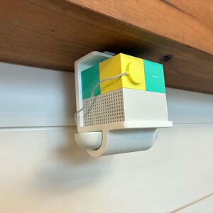 May include: A multi-colored, geometric-shaped device mounted on a white wall. The device has a white base with a perforated front, and a top section with yellow, teal, and green blocks. A yellow cord connects the blocks. The device is mounted under a wooden shelf.