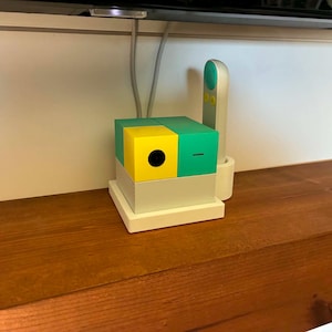 May include: A modern, geometric device with a yellow and green cube design, featuring a black circular element and a white base. A white remote with a green button is also present. The device sits on a wooden surface.