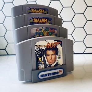 Può includere: Quattro cartucce di gioco per Nintendo 64 impilate l'una sull'altra. Le cartucce sono grigie e hanno etichette colorate. Le etichette includono i titoli "Super Smash Bros", "Super Smash Bros", "Super Mario 64" e "GoldenEye 007".