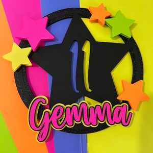 May include: A black star-shaped cake topper with the number 11 in yellow, surrounded by a glittery black circle. The name "Gemma" is in pink with a black outline. Five colorful stars in pink, orange, and green are scattered around the star.