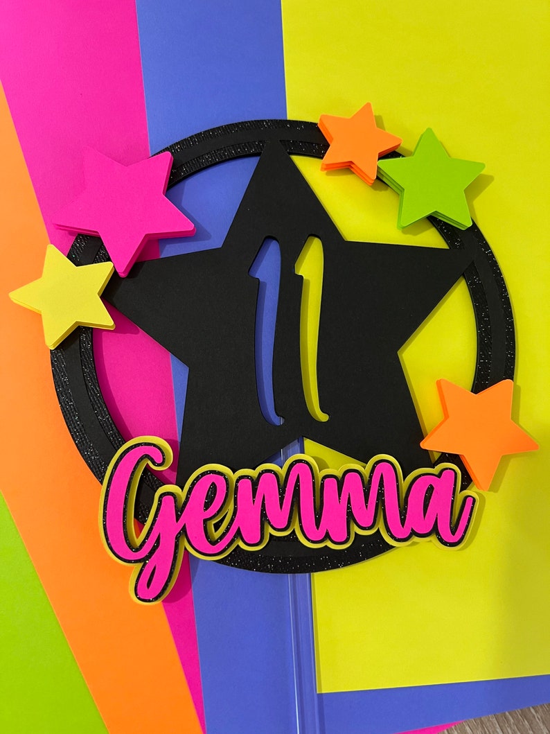 May include: A black star-shaped cake topper with the number 11 in yellow, surrounded by a glittery black circle. The name "Gemma" is in pink with a black outline. Five colorful stars in pink, orange, and green are scattered around the star.