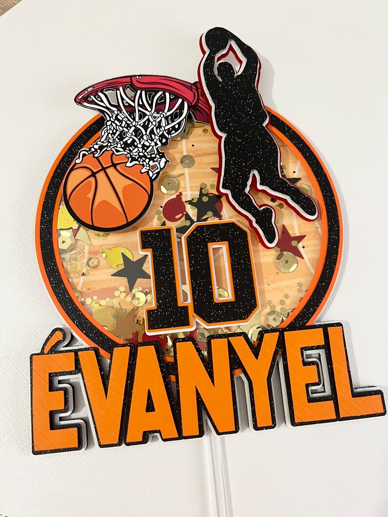 May include: A basketball-themed cake topper featuring a basketball, hoop, and a player shooting a basket. The topper also includes the number 10 and the name "EVANYEL" in orange and black.