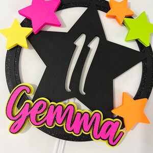 May include: A black star-shaped cake topper with the number 11 in white. The topper is surrounded by a black glitter ring and colorful foam stars in pink, yellow, and orange. The name "Gemma" is written in pink with a yellow outline.