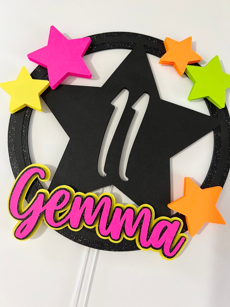 May include: A black star-shaped cake topper with the number 11 in white. The topper is surrounded by a black glitter ring and colorful foam stars in pink, yellow, and orange. The name "Gemma" is written in pink with a yellow outline.