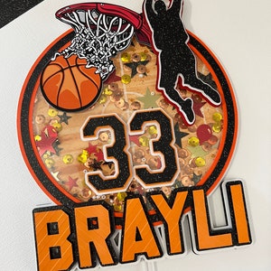 May include: Orange and black basketball themed cake topper with a basketball, hoop, player silhouette, and the number 33. The name "BRAYLI" is written in orange letters with black outlines.