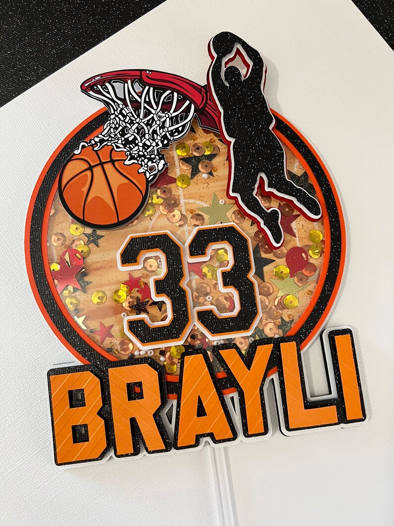 May include: Orange and black basketball themed cake topper with a basketball, hoop, player silhouette, and the number 33. The name "BRAYLI" is written in orange letters with black outlines.