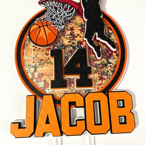 May include: Orange and black basketball themed cake topper with a basketball going through a hoop, a player silhouette, the number 14, and the name Jacob.