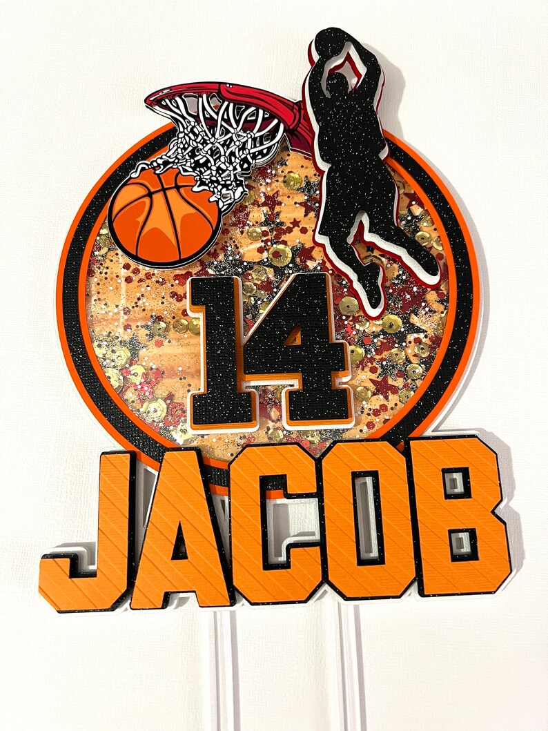 May include: Orange and black basketball themed cake topper with a basketball going through a hoop, a player silhouette, the number 14, and the name Jacob.