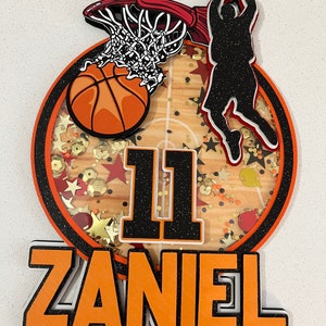 May include: Orange and black basketball themed cake topper with the number 11 and the name ZANIEL. The topper features a basketball, a basketball hoop, and a silhouette of a basketball player.