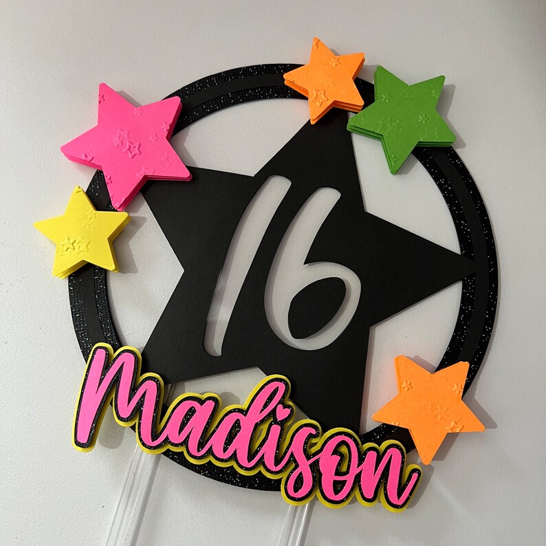 May include: A black star-shaped cake topper with the number 16 cut out. The topper is surrounded by a glittery black ring and colorful star accents in pink, yellow, orange, and green. The name "Madison" is written in pink with a yellow outline.