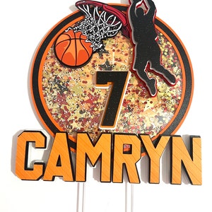 May include: Orange and black basketball themed cake topper with a basketball, hoop, player, and the number 7. The name Camryn is written in orange and black letters.