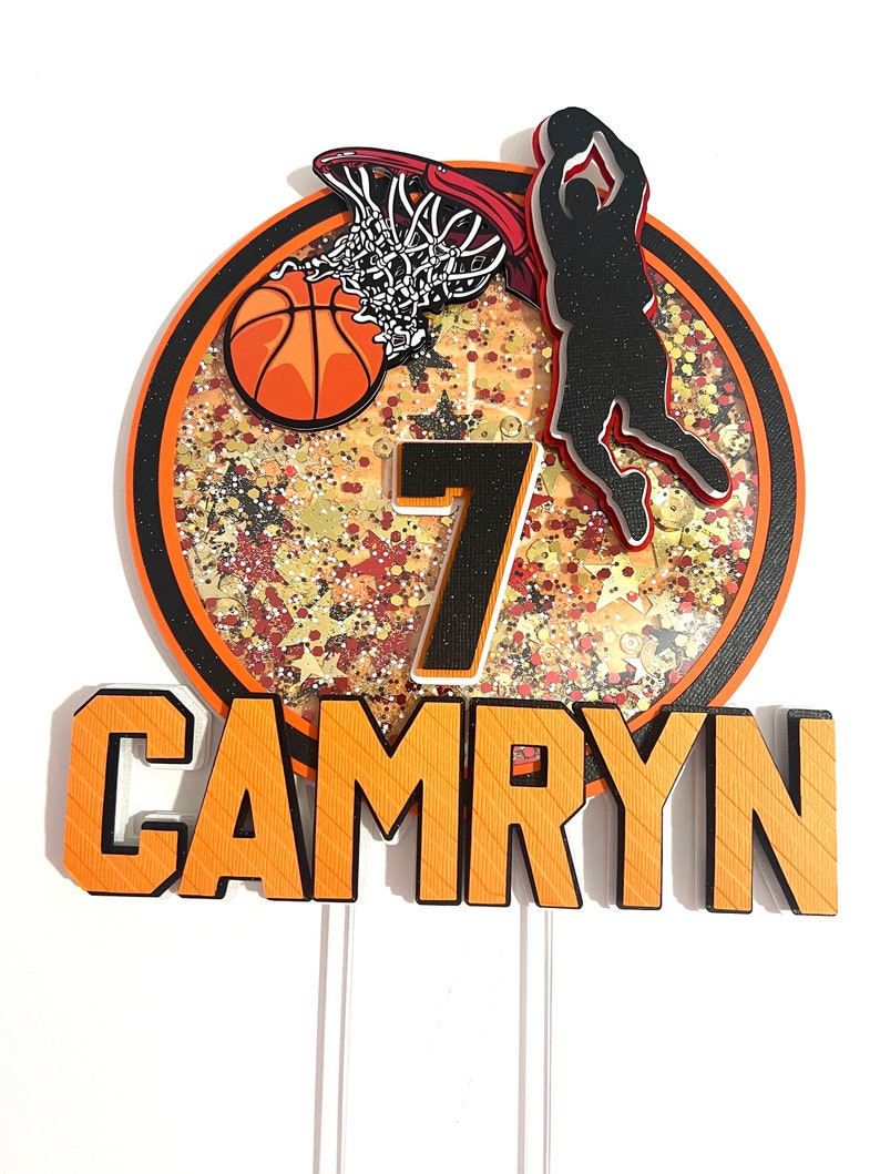 May include: Orange and black basketball themed cake topper with a basketball, hoop, player, and the number 7. The name Camryn is written in orange and black letters.