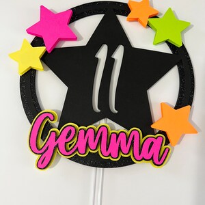 May include: A black star-shaped cake topper with the number 11 cut out. The topper has a glittery black border and is surrounded by colorful star accents in pink, yellow, orange, and green. The name "Gemma" is written in pink with a yellow outline.