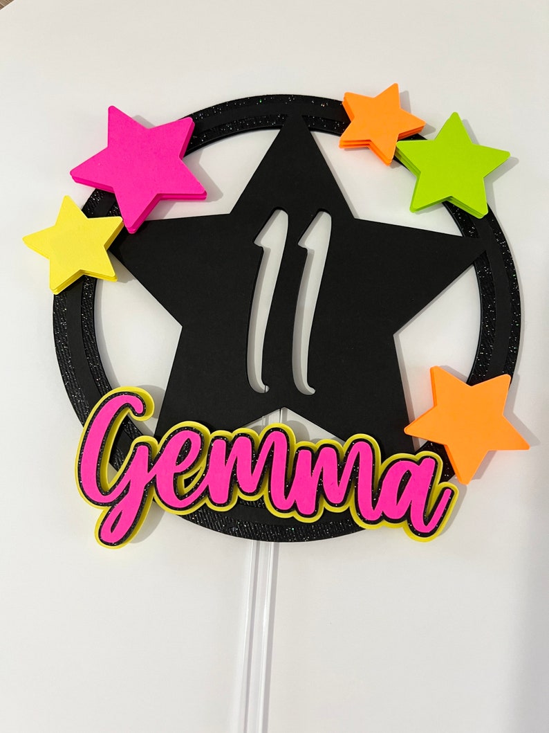 May include: A black star-shaped cake topper with the number 11 cut out. The topper has a glittery black border and is surrounded by colorful star accents in pink, yellow, orange, and green. The name "Gemma" is written in pink with a yellow outline.
