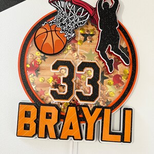 May include: A basketball themed cake topper featuring a basketball, hoop, and player silhouette. The number 33 and the name "BRAYLI" are also included.