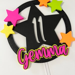 May include: A black star-shaped cake topper with the number 11 cut out. The topper is surrounded by a black glittery ring and accented with neon pink, orange, and yellow stars. The name "Gemma" is written in pink with a yellow outline.