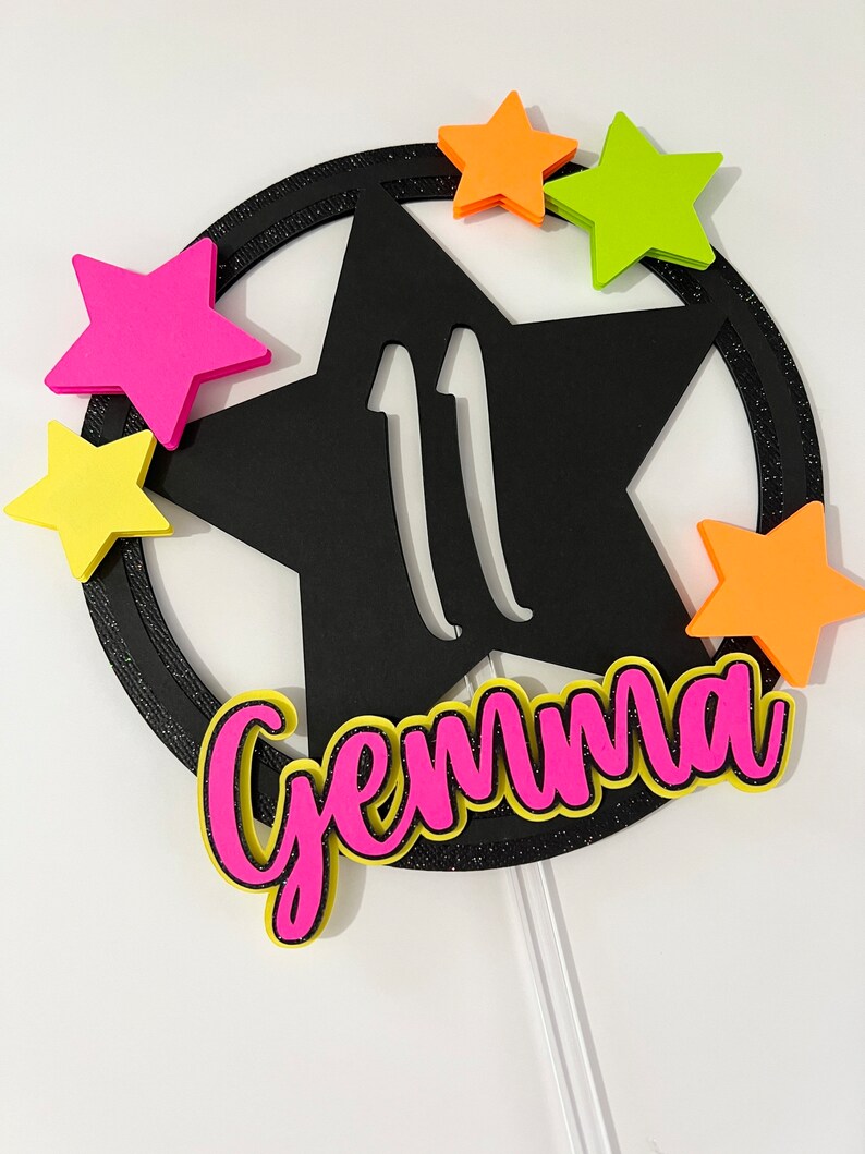 May include: A black star-shaped cake topper with the number 11 cut out. The topper is surrounded by a black glittery ring and accented with neon pink, orange, and yellow stars. The name "Gemma" is written in pink with a yellow outline.