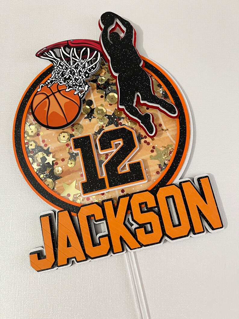 May include: A black and orange basketball themed cake topper with a basketball player shooting a hoop, a basketball, the number 12, and the name JACKSON.