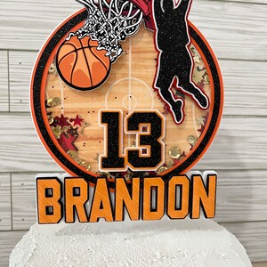 May include: A basketball themed cake topper featuring a basketball player shooting a hoop, a basketball, and the number 13. The topper also includes the name "BRANDON" in orange letters.