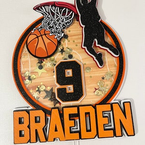 May include: Orange and black basketball themed cake topper with a basketball going through a hoop, a silhouette of a player dunking, the number 9, and the name "Braeden".